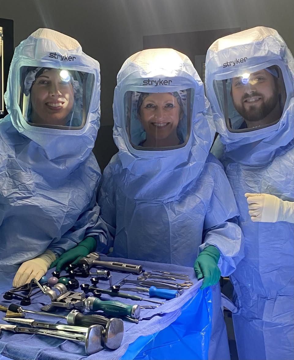 Dr. MacNeille and two members of his surgical team in sterile surgical hoods and gowns, standing behind a table of joint replacement instruments.